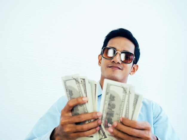 Portrait of happy rich asian young businessman in casual blue\
shirt and wearing sunglasses enjoying while holding and counting\
dollars money banknotes on white wall background