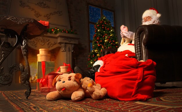 Portrait of happy Santa Claus sitting at his room at home near Christmas tree and reading Christmas letter or wish list.