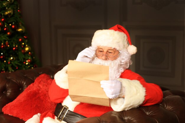 Portrait of happy Santa Claus sitting at his room at home near Christmas tree and reading Christmas letter or wish list.