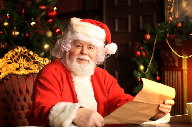 Portrait of happy Santa Claus sitting at his room at home near Christmas tree and reading Christmas letter or wish list.