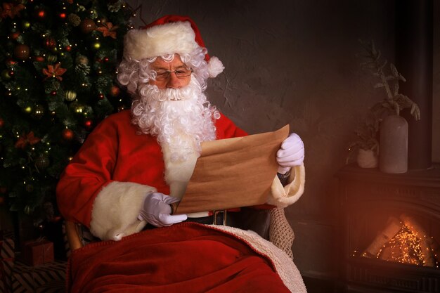 Portrait of happy Santa Claus sitting at his room at home near fireplace and reading Christmas letter or wish list.