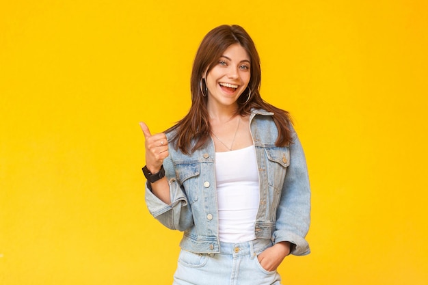 Portrait of happy satisfied beautiful brunette young woman with makeup in denim casual style standing, thumbs up and looking at camera excited. indoor studio shot, isolated on yellow background.