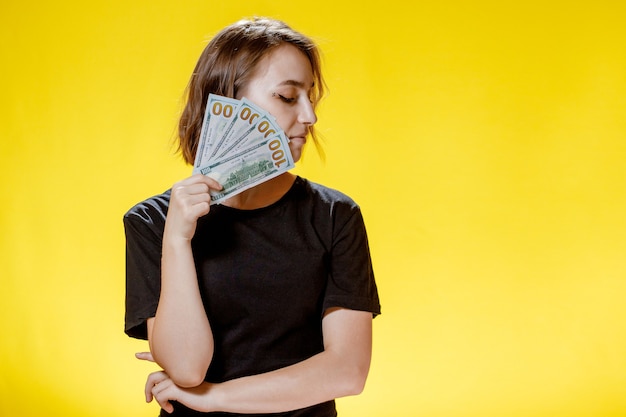 Портрет счастливой довольной девушки, держащей кучу денежных банкнот и смотрящей в камеру на желтом фоне.