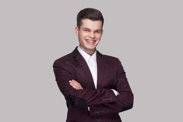 Portrait of happy satisfied handsome young man in violet suit and white shirt, standing, crossed hands and looking at camera with toothy smile. indoor studio shot, isolated on grey background.