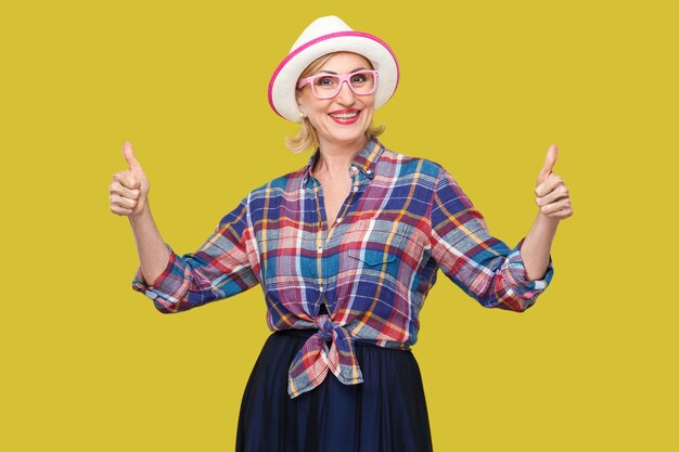 Portrait of happy satisfied modern stylish mature woman in casual style, hat and eyeglasses standing, thumbs up, looking at camera with toothy smile. indoor studio shot isolated on yellow background.