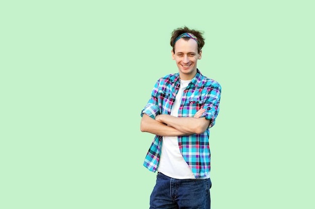 Portrait of happy satisfied young man in casual blue checkered shirt and headband standing, looking at camera with croseed arms and toothy smile. indoor studio shot, isolated on light green background