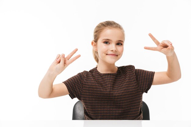 Portrait of happy school girl smiling and showing peace sign while sitting at desk in class isolated over white wall