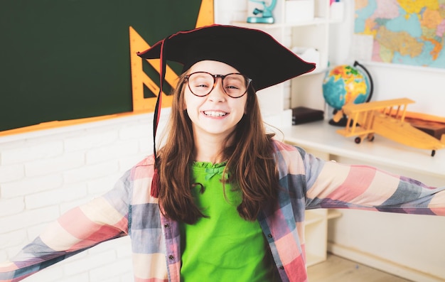 Portrait of happy schoolgirl with graduation hat in classroom