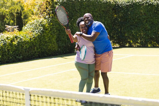 Portrait of happy senior african american couple with rackets embracing on sunny grass tennis court. Senior lifestyle, retirement, sport, summer, fitness, hobbies and leisure activities.