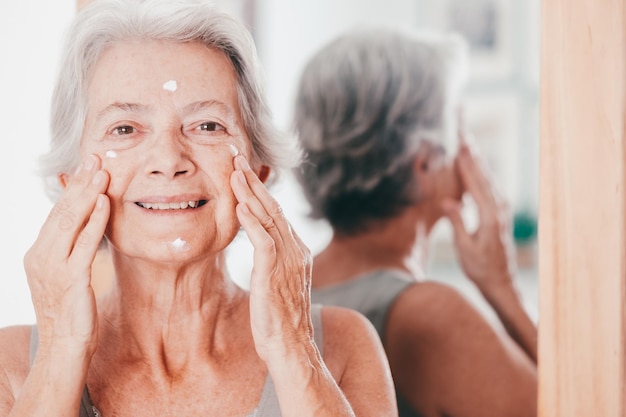 Portrait of happy senior beautiful woman applies anti aging cream on wrinkled face elderly smiling