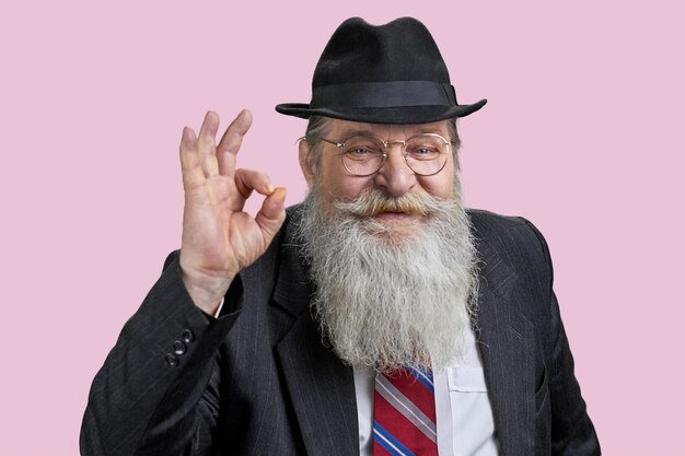 Portrait of happy senior man showing okay gesture Long beard and black suit Isolated on pink