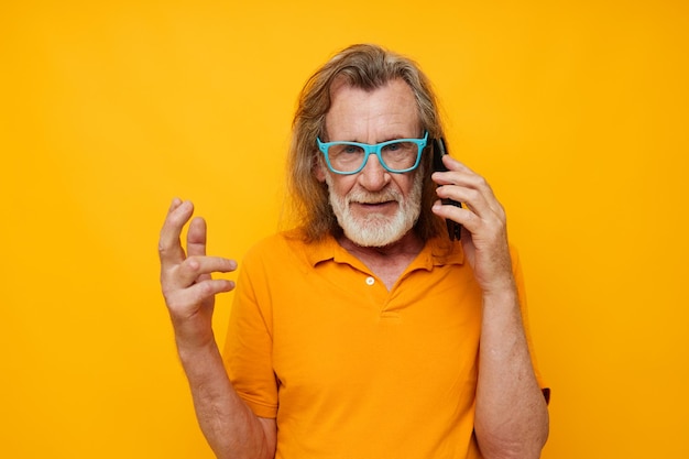 Portrait of happy senior man wearing blue glasses yellow shirt talking on the phone yellow background