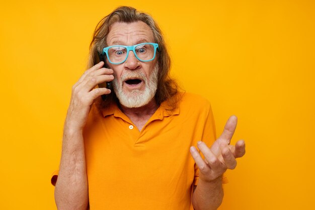 Portrait of happy senior man wearing blue glasses yellow shirt talking on the phone yellow background