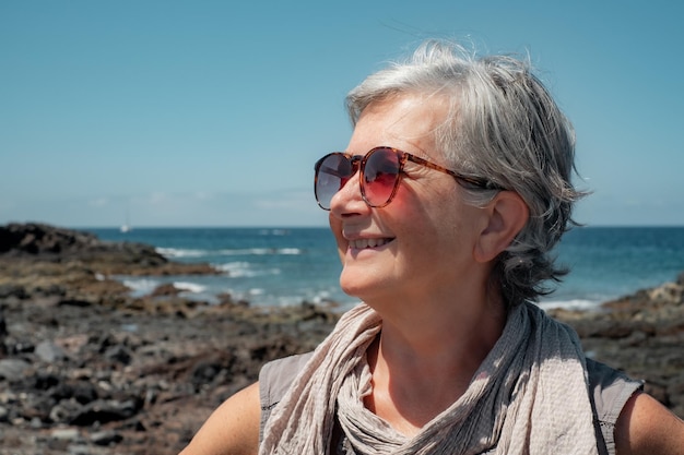 Portrait of happy senior mature woman with sunglasses enjoying sea vacation standing at the beach