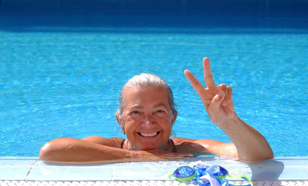 Portrait of an happy senior sporty woman enjoying swimm into the swimming pool doing ok gesture with hand  - active retiree enjoying swimming on a sunny day