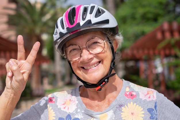 Foto ritratto di donna anziana felice che indossa il casco da ciclismo all'aperto guardando la fotocamera che gesturing il segno della vittoria signora anziana sorridente