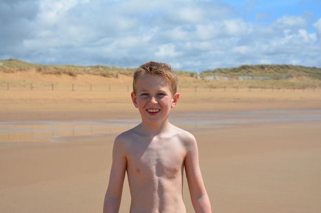 Photo portrait of happy shirtless boy standing at beach against sky on sunny day