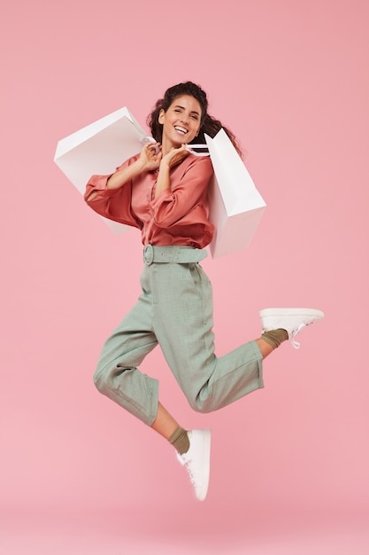 Portrait of happy shopaholic enjoying her shopping she jumping up with shopping bags against the pink background