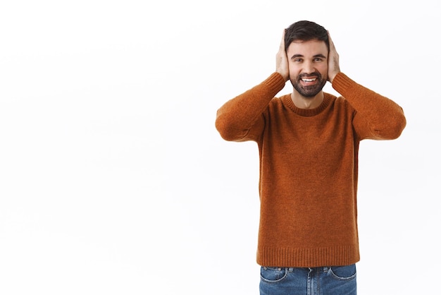 Portrait of happy sincere bearded man cover ears and smiling pretend not hear unwilling listen look camera delighted and upbeat standing enthusiastic in brown sweatshirt over white background