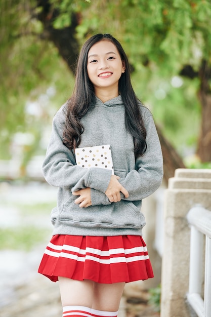 Portrait of happy smiling Asian schoolgirl holding journal or diary and  when standing in park