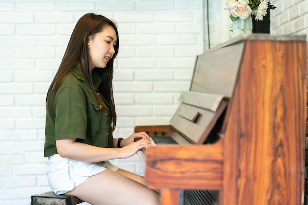 Портрет счастливой улыбающейся красивой азиатки в коричнево-зеленом платье, стильный хипстер с игрой на старом деревянном пианино в винтажном классическом стиле