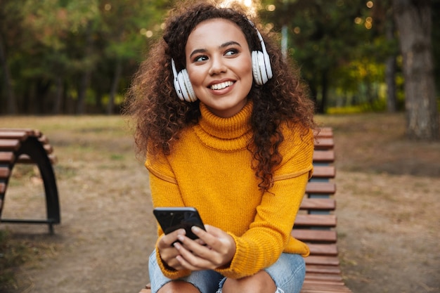 幸せな笑顔の美しい若い巻き毛の女性の肖像画は、携帯電話を使用してヘッドフォンで音楽を聴いて屋外公園のベンチに座っています。