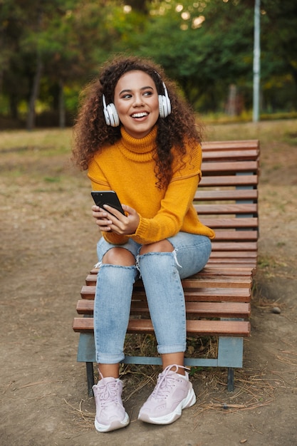 幸せな笑顔の美しい若い巻き毛の女性の肖像画は、携帯電話を使用してヘッドフォンで音楽を聴いて屋外公園のベンチに座っています。