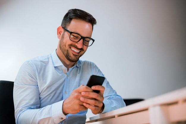 사무실에 앉아있는 동안 스마트 폰을 사용하는 안경에 행복 웃는 사업가의 초상화.