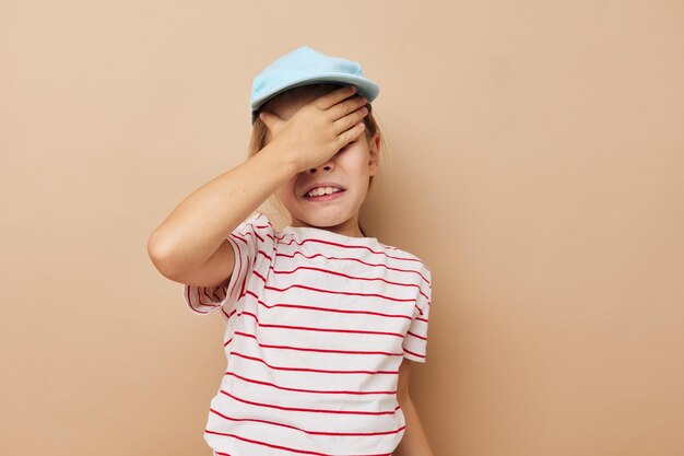 青い帽子の縞模様のTシャツの感情ベージュの背景で幸せな笑顔の子供の女の子の肖像画