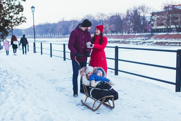 Portrait of happy smiling couple, looking at each other while walking with their kids. Two cute toddlers wearing warm winter clothes sitting in wooden sleigh. Winter city on background