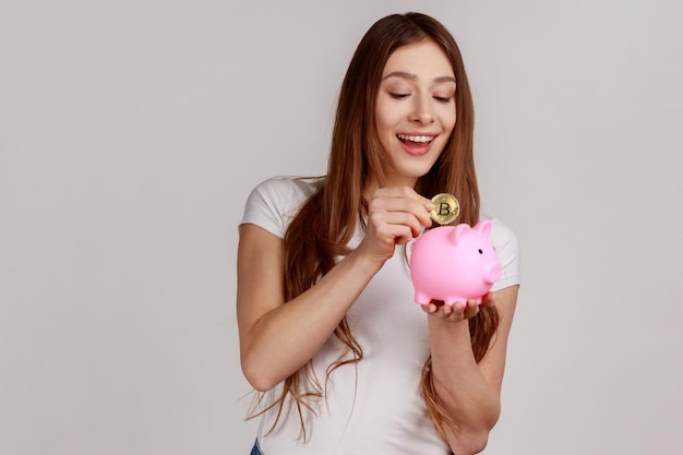 Portrait of happy smiling dark haired woman investing in bitcoins, putting golden crypto coin into piggy bank, wearing white T-shirt. Indoor studio shot isolated on gray background.