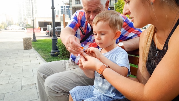 Portrait of happy smiling family with little toddler boy, young mather and grandfather sitting on bench at park and playing with small plastic toy