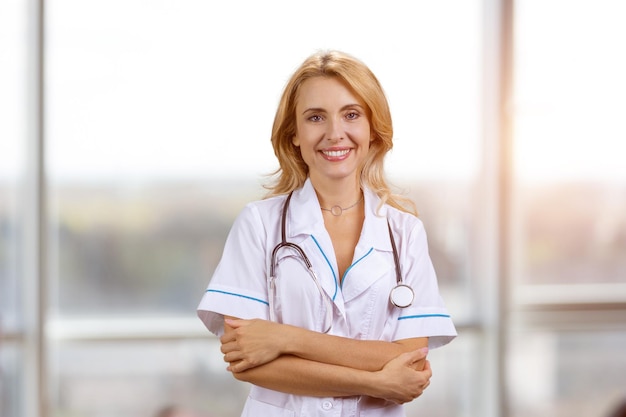 Portrait of happy smiling female doctor with folded arms indoor window in the background