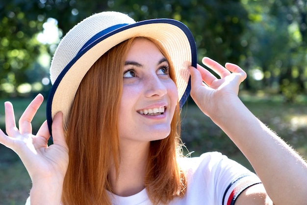 Portrait of happy smiling girl with red hair and in yellow hat outdoors in summer park.
