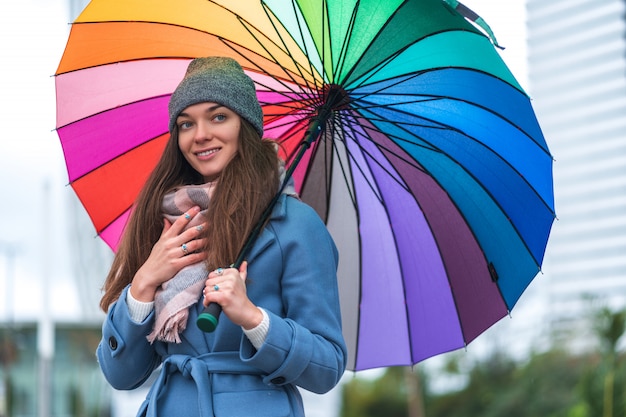 秋の雨の日と雨の天候の間に明るい色のレインボーパラソルと暖かい服装で幸せな笑顔のうれしそうな屈託のない女の肖像画。雨からの保護