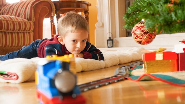 Portrait of happy smiling little boy lying on wooden floor and looking on riding train on railroad. Child receiving presents and toy on New Year or Xmas