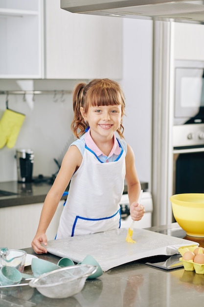 Portrait of happy smiling little girl using silicone brush when applying melted butter on baking sheet