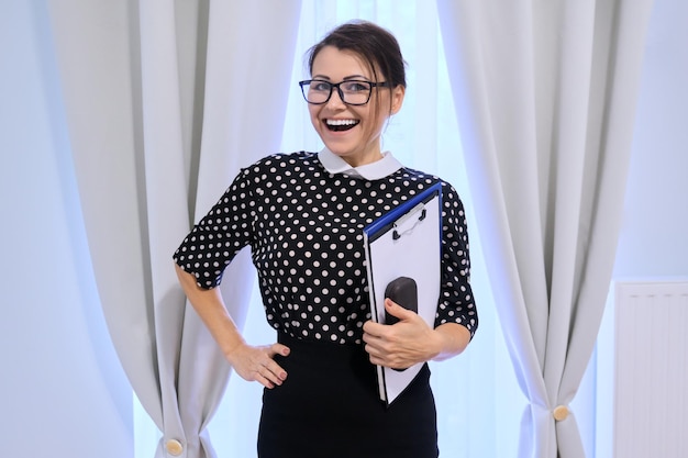 Portrait of happy smiling mature business woman in glasses with papers, female looking at camera, inside near the window with curtains