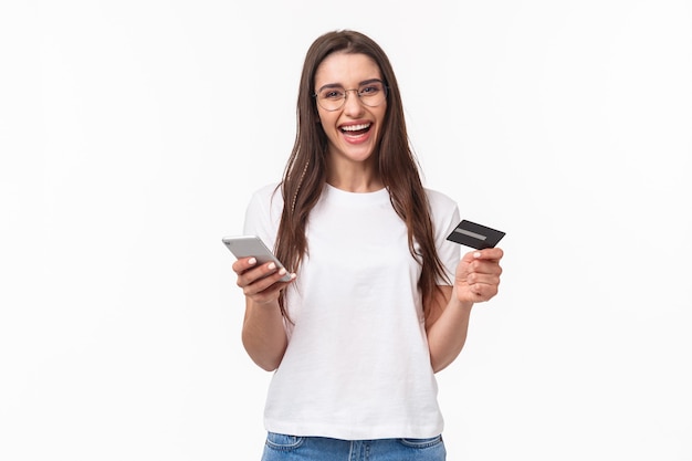 Portrait of happy smiling pretty woman in glasses holding credit card, mobile phone, laughing at camera easily paying for her purchase online store