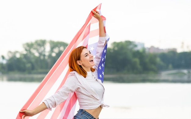 Portrait of happy smiling red haired girl with USA national flag in her hands. Positive young woman celebrating United States independence day.