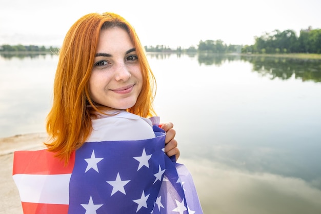 Portrait of happy smiling red haired girl with USA national flag on her shoulders Positive young woman celebrating United States independence day International day of democracy concept