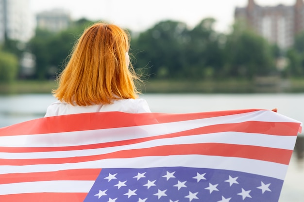 Foto ritratto della ragazza dai capelli rossi sorridente felice con la bandiera nazionale di usa sulle sue spalle