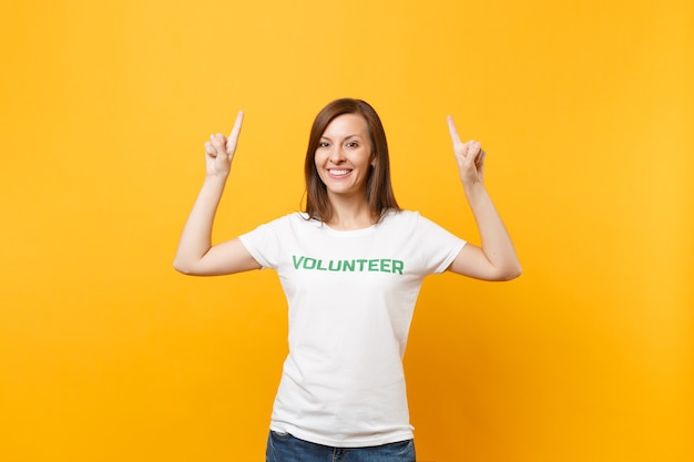 Portrait of happy smiling satisfied woman in white t-shirt with written inscription green title volunteer isolated on yellow background. voluntary free assistance help, charity grace work concept