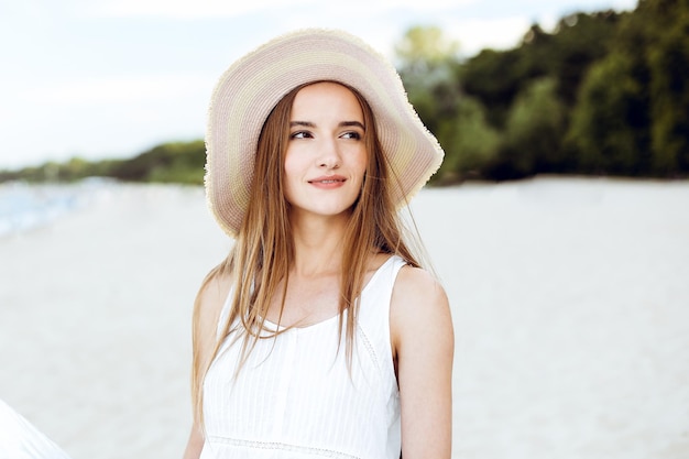 Portrait of a happy smiling woman in free happiness bliss on ocean beach standing with a hat. A female model in a white summer dress enjoying nature during travel holidays vacation outdoors