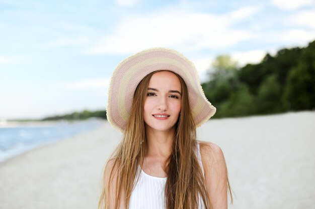 Portrait of a happy smiling woman in free happiness bliss on ocean beach standing with a hat. A female model in a white summer dress enjoying nature during travel holidays vacation outdoors