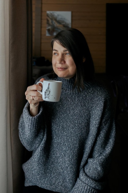 Портрет счастливой улыбающейся женщины девушки с чашечкой кофе женщины, пьющей чай дома у окна