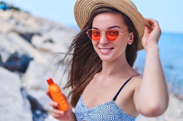 Portrait of a happy smiling woman in a swimsuit, straw hat and red sunglasses with a bottle of sun block cream while sunbathing by the sea in the summertime