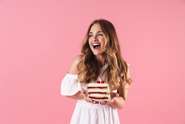 Ritratto di una donna sorridente felice che indossa un abito bianco che guarda da parte e tiene in mano un pezzo di torta isolato su un muro rosa