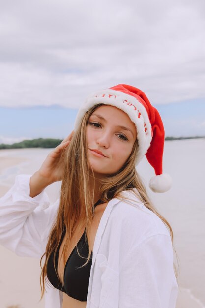 Portrait of happy smiling woman with tropical background at white sand beach female is wearing Santa hat white shirt and bikini