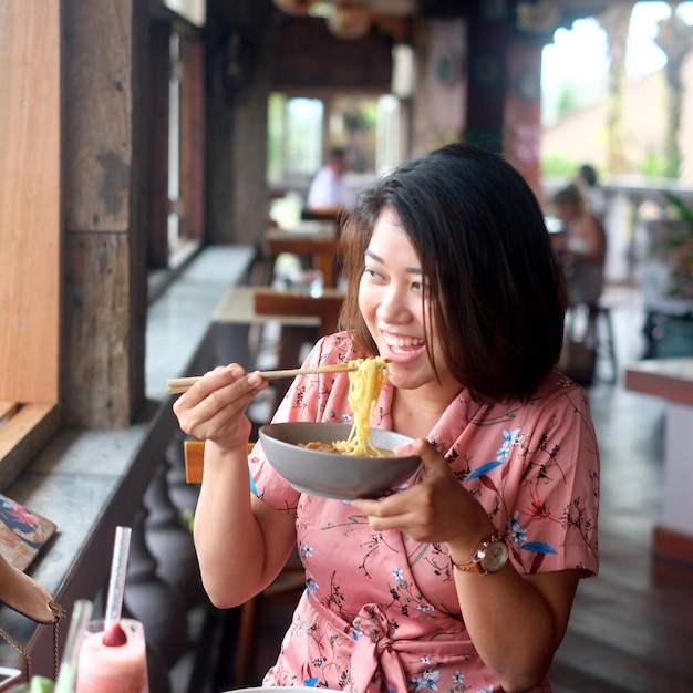 Portrait of happy smiling young Asian woman eating Asian noodles